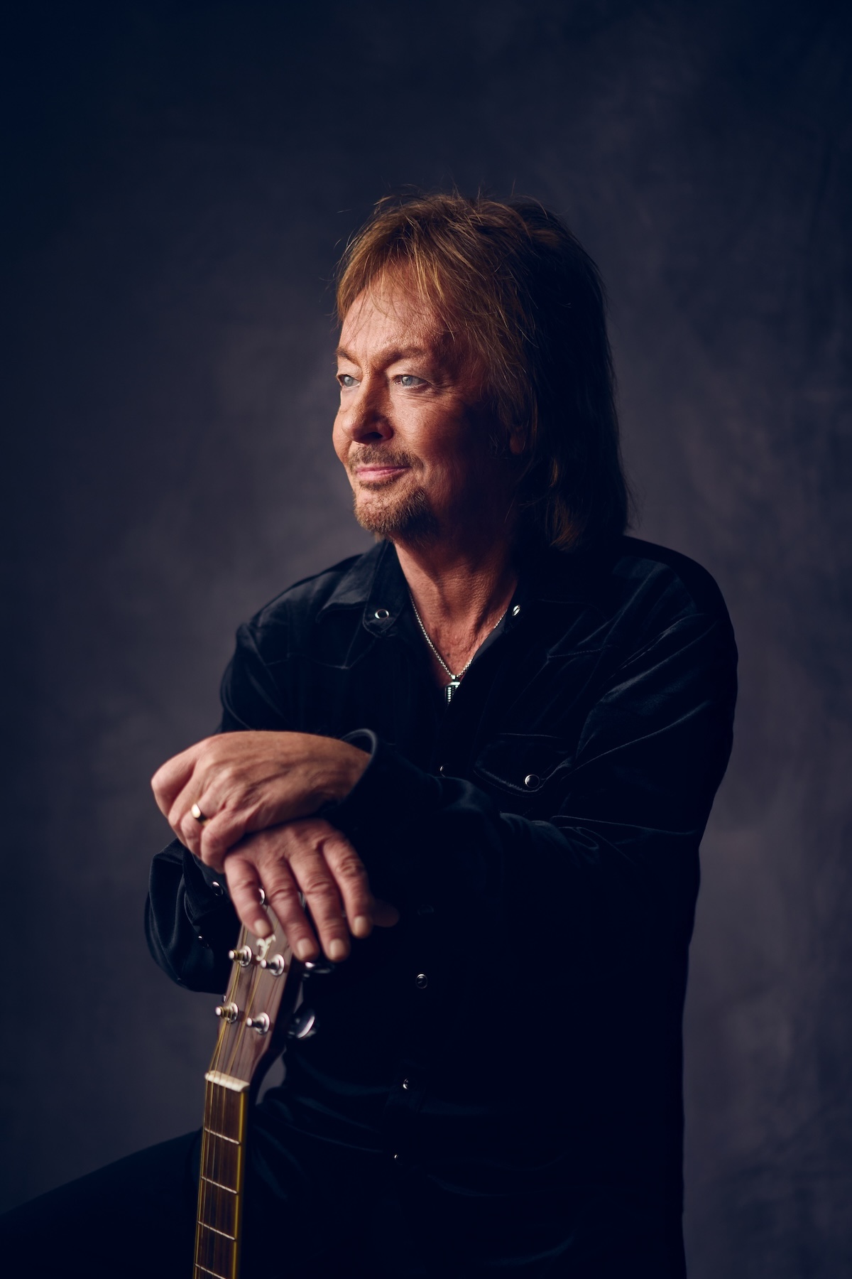 Chris Norman stands, looks to the side and his hands rest on a standing guitar